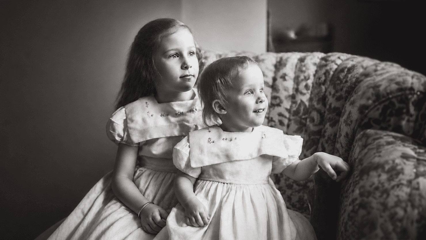 Two young girls in matching embroidered dresses sit on a floral sofa, black and white.